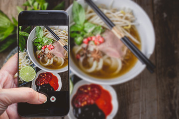 Wall Mural - Taking a photo by Finger Pressing on Smartphone for Photograph Pho Vietnamse Noodle Soup on Wooden Background with Copy Space. Image for Food Advertise Concept