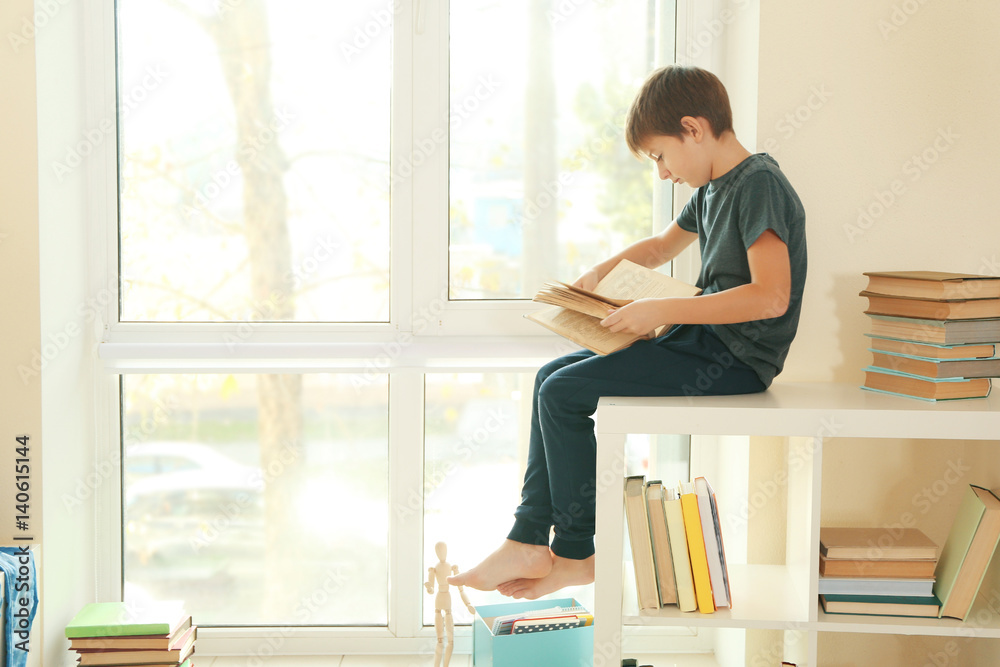 Cute schoolboy sitting beside window and reading book Stock Photo ...