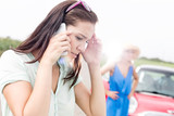 Fototapeta  - Worried woman using mobile phone while friend standing by broken down car on sunny day