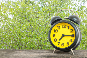 Closeup black and yellow alarm clock for decorate show a quarter past seven or 7:15 a.m. on old brown wood desk on green leaves in the park textured background