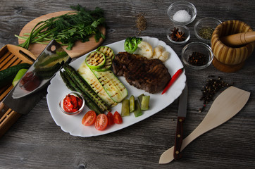 Wall Mural - Grilled beef grilled with asparagus, zucchini, lobi, broccoli, tomato, chili and red sauce on a white plate, and other vegetables in wood background