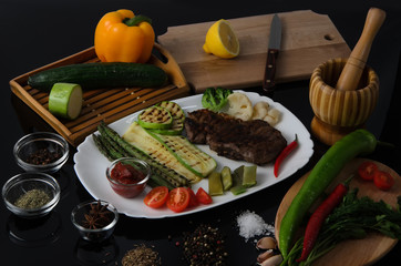 Wall Mural - Grilled beef grilled steak with asparagus, zucchini, lobi, broccoli, tomato, chili and red sauce on a white plate, on a black glossy background