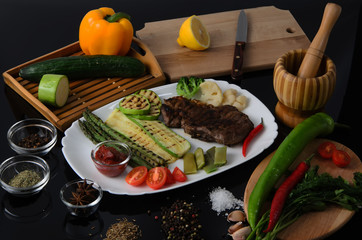 Wall Mural - Grilled beef grilled steak with asparagus, zucchini, lobi, broccoli, tomato, chili and red sauce on a white plate, on a black glossy background