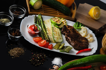 Wall Mural - Grilled beef grilled steak with asparagus, zucchini, lobi, broccoli, tomato, chili and red sauce on a white plate, on a black glossy background