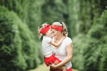 Woman holds little laughing girl in her arms standing among green trees