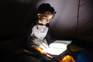 Teen boy sitting in a camping tent wrapped in a sleeping bag reading a book with flashlight on at night