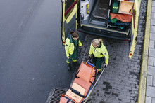 High Angle View Of Female Paramedic Standing With Colleague Pushing Stretcher Outside Ambulance On Road
