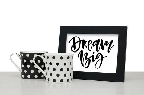 Dream big. Handwritten text, inspirational quote. Modern calligraphy. Black wooden frame. Black and white coffee Cup on the table