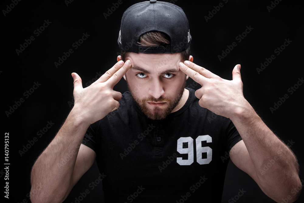 portrait of handsome young man in black shirt and cap on black background. rapper - obrazy, fototapety, plakaty 
