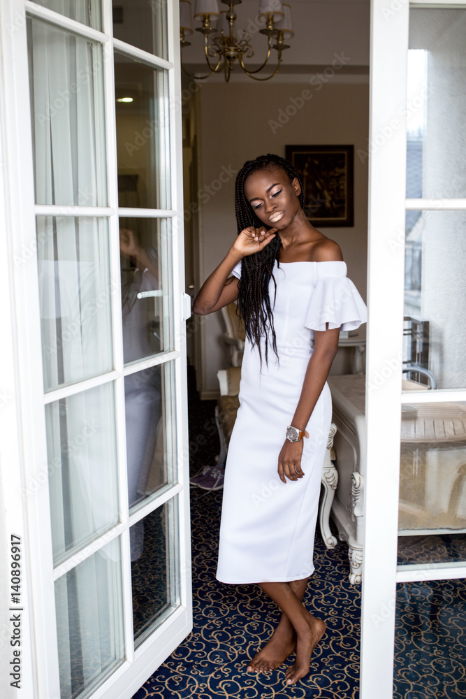 Pretty young girl with dark shiny skin standing near the french window in white  dress. Afro american woman Stock Photo | Adobe Stock