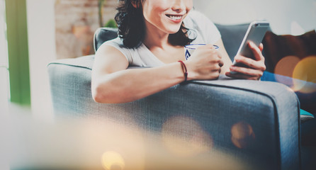 Young smiling Asian woman spending rest time at home on sofa, holding ceramic cup at hand and using smartphone for video conversation with friends.Blurred background, flares effect.Wide.