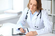 © rogerphoto - Young female doctor filling up medical form while sitting at the table near window  in hospital