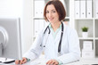 © rogerphoto - Young brunette female doctor sitting at a desk and working on the computer at the hospital office.  Health care, insurance and help concept. Physician ready to examine patient