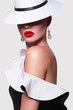 A girl in a white hat and in black and white attire with a shaft in the studio on a white background. A girl with red lips and smooth skin stands sideways.