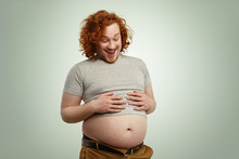 Indoor Shot Of Funny Overweight Young Caucasian Male Wearing Undersized Grey T-shirt, Holding Hands On His Stomach, Looking At His Big Belly Hanging Out Of Pants After Eating Fast Food On Dinner