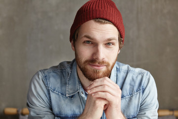 Isolated portrait of handsome young bearded European male sitting against concrete wall background, keeping hands clasped, looking at camera with happy smile, wearing stylish hat and denim jacket