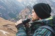 A hipster man with a beard in a hat, a jacket, and a backpack in the mountains holds binoculars, adventure, tourism, tracking