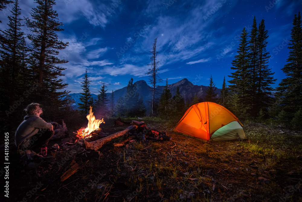 Man looking up at the stars next to campfire and tent at night - obrazy, fototapety, plakaty 