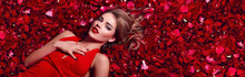 Valentines Day. Loving Girl. The Girl In A Red Dress Lying On The Floor In The Petals Of Red Roses. Background Of Red Rose Petals. Red Lipstick On The Lips From The Beautiful Girl.