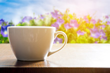 Hot Coffee In White Cup On Wooden Table In The Garden. Purple Morning Glory Flower In The Background And Sunlight Shining From The Right.