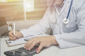 young medical doctor caucasian healthcare professional wearing a white coat with stethoscope in hospital ,doctor's office calculates on an electronic calculator,selective focus,vintage color