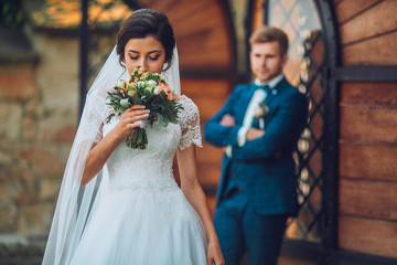 Handsome man groom hugging pretty girl or cute bride with beautiful brunette hair in white wedding dress outdoors at ancient building door on grey background