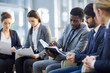 © pressmaster - Multi-ethnic group of  business people sitting in row in modern glass hall, focus on African-American businessman reading handout materials and making notes in book