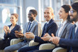 © pressmaster - Multi-ethnic group of smiling business people sitting in row in modern glass hall and clapping, focus on young cheerful  businessman in center