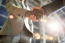 Low Angle Closeup Shot Of Two Business Partners In Handshake: Unrecognizable African -American Businessman Shaking Hands With Caucasian Colleague In Hall Of Modern Glass Office Building At Night Time