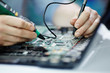 © pressmaster - Closeup shot of hands testing electric current in circuit board of disassembled laptop using multimeter tool on table in maintenance shop