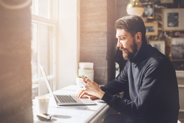 Young man drinking coffee in cafe and using laptop, Portrait of attractive man checking new messages on smartphone while working in modern cafe