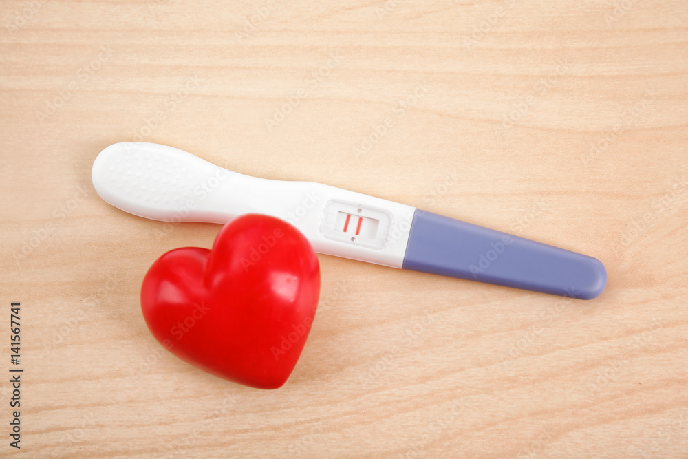 Pregnancy test and red plastic heart on wooden background