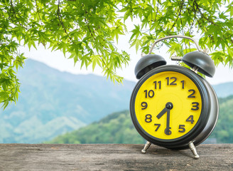 Closeup black and yellow alarm clock for decorate show half past seven or 7:30 a.m. on blurred leaves and mountain view background