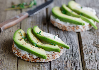 Close up of two toasts of grain bread with soft white cheese and chopped fresh avocado on old wooden gray boards .