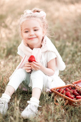 Poster - Smiling baby girl 3-4 year old eating strawberries sitting outdoors. Looking at camera. Summer time.