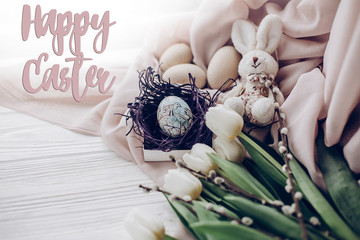 happy easter text greeting card sign on stylish easter eggs with chick ornaments in nest and bunny rabbit and tulips and willow buds on rustic white wooden background. space for text