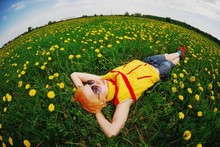 Art Portrait With The Fisheye Effect Of A Young Pretty Blonde Woman With Short Hair In Yellow-red Vest, Wearing Sunglasses, Relaxing On Meadow Of Yellow Flowers Of Dandelions.