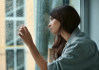 Depressed young woman near window at home