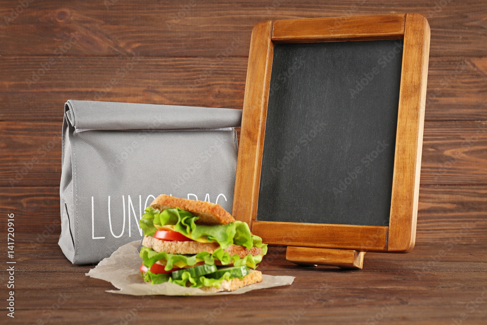 Lunch bag, small blackboard and sandwich on wooden background