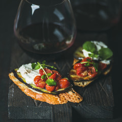 Poster - Wine and snack set. Brushetta with roasted eggplant, cherry tomatoes, garlic, cream cheese, arugula and glass of red wine on wooden board over dark background, selective focus, square crop. Slow food