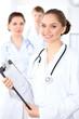© rogerphoto - Happy female doctor keeping medical clipboard while medical staff are at the background. Successful team at health care and medicine concept