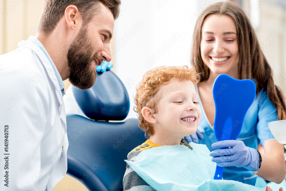 Young boy looking at the mirror with toothy smile sitting on the chair with dentist and assistant at the dental office - obrazy, fototapety, plakaty 