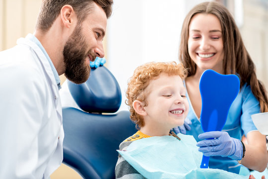 young boy looking at the mirror with toothy smile sitting on the chair with dentist and assistant at