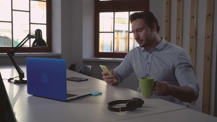 Poster - adult businessman sitting at the desk in office. Smiling handsome man using smartphone drinking coffee looking on screen laptop. Male wearing in casual shirt typing on computer in room with windows.
