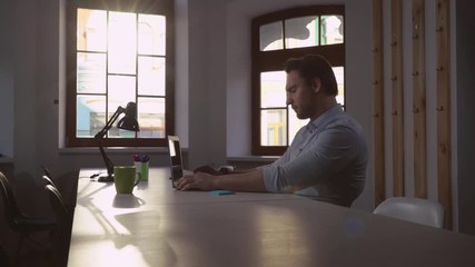 Poster - Businessman working in the office in sunny day. Handsome adult man typing on computer in office. Concentrated male sitting at the big wooden table looking screen laptop.