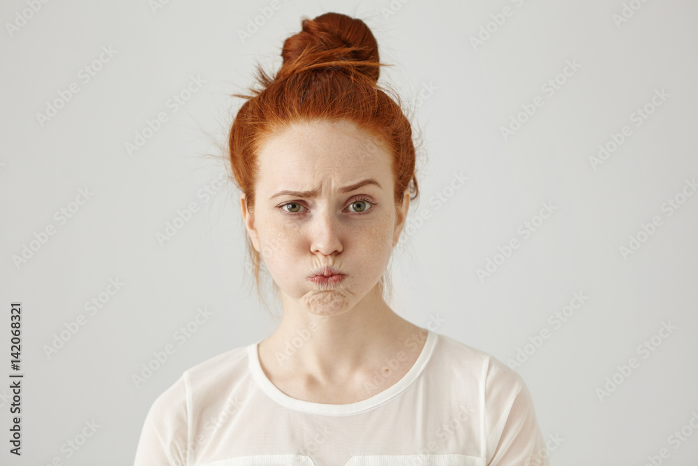 Headshot of attractive funny young female with ginger hair dressed in white blouse feeling displeased or uncomfortable with something, blowing cheeks and frowning. Human face expressions and emotions - obrazy, fototapety, plakaty 