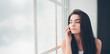 © estradaanton - A young woman with black hair in a white T-shirt at a window flooded with sunlight uses a smartphone, communicates and searches for information on the Internet or social networks