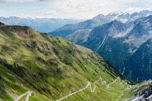 Alp Road Surrounded By Green Hills And Blue Alp High Mountains On Background. Steep Descent Of Passo Dello Stelvio In Stelvio Natural Park, Tyrol, Italy.