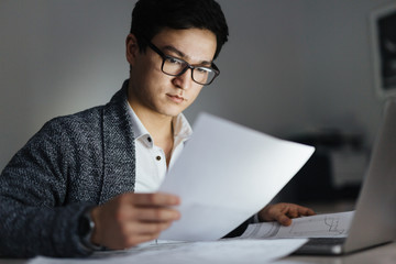 Portrait of busy Asian man wearing glasses and casual wear sorting documentation and working with laptop in dark room late at night, his face lit up by screen