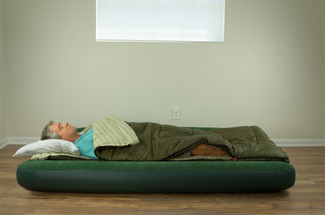 Man comfortably sleeping on blow up air mattress bed in sleeping bag with clean walls and window in the background.